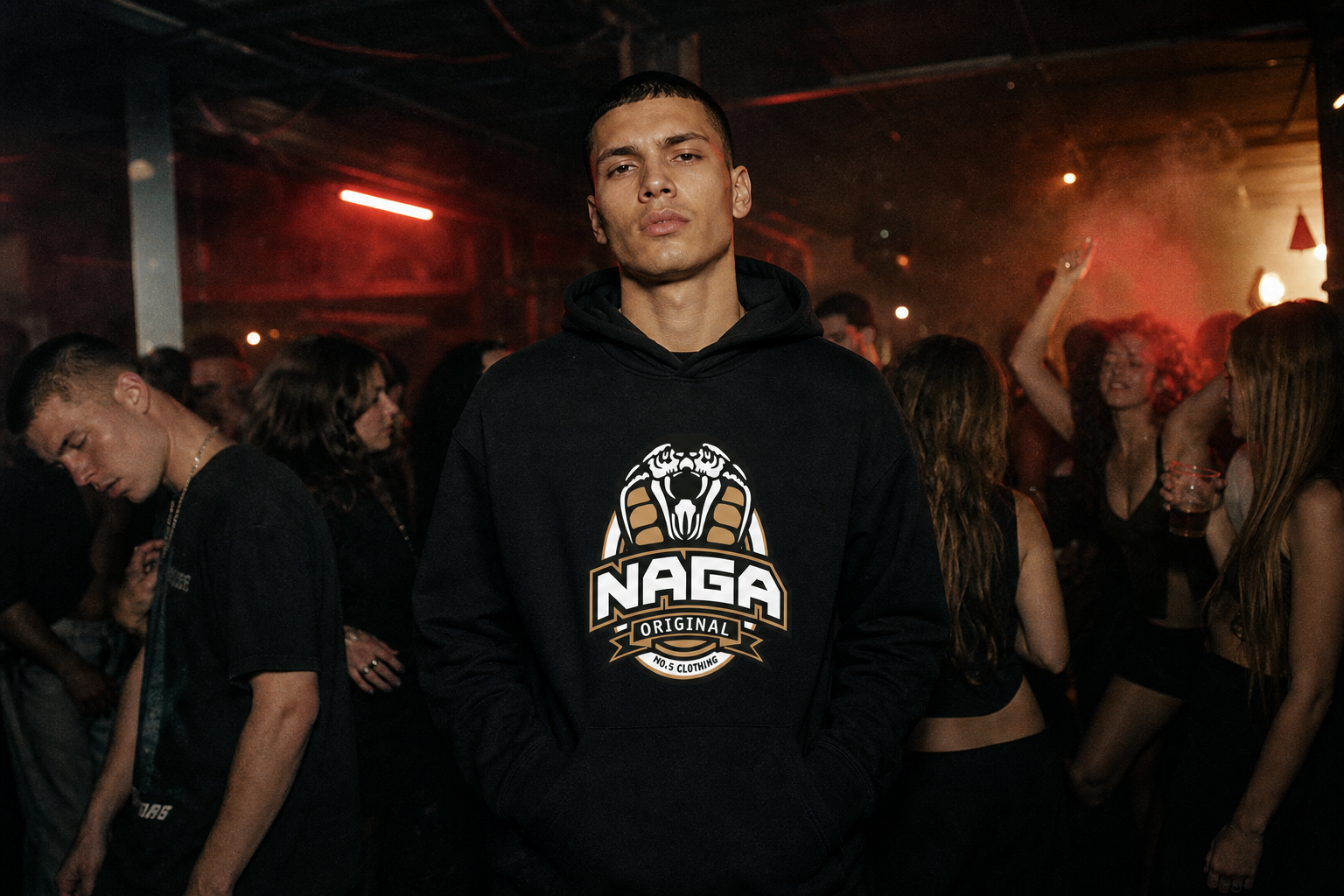 Model wearing a black Naga Original hoodie with cobra chest logo in a dimly lit crowd